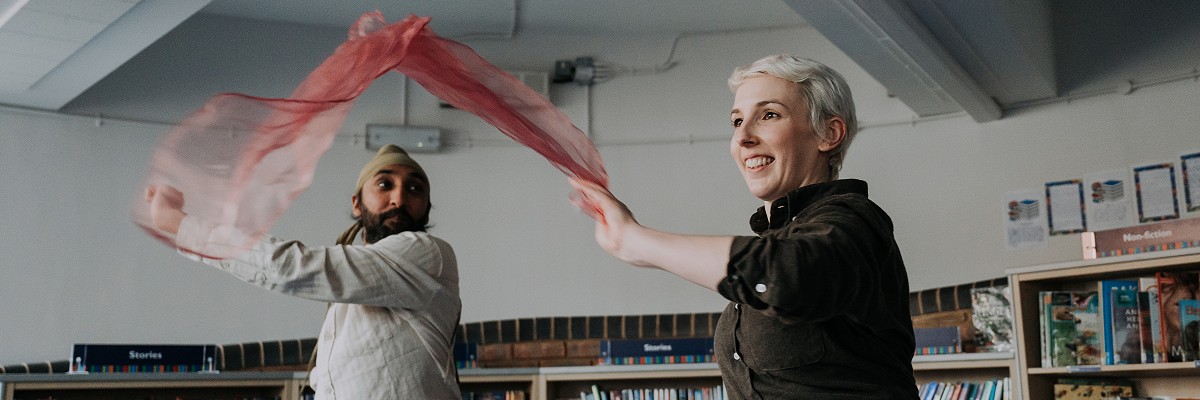 Two dancers throw colourful scarves forward and try to catch them. They are dancing in the children's section of a library.
