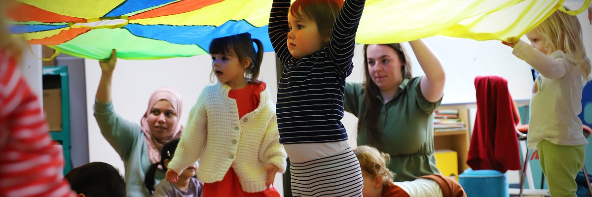 A group of children and their grown-ups dance under a large multi-coloured parachute, as part of the research for the SEED performance